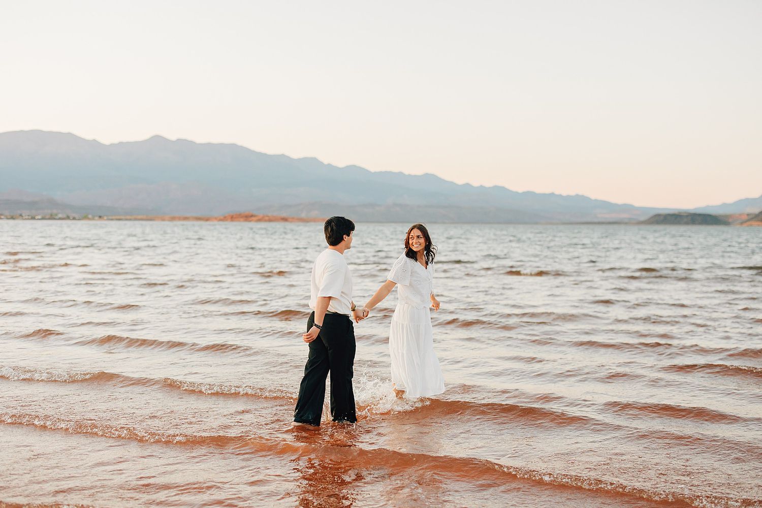 Couple holding hands standing in shallow ocean water at sunset with mountains in the background.