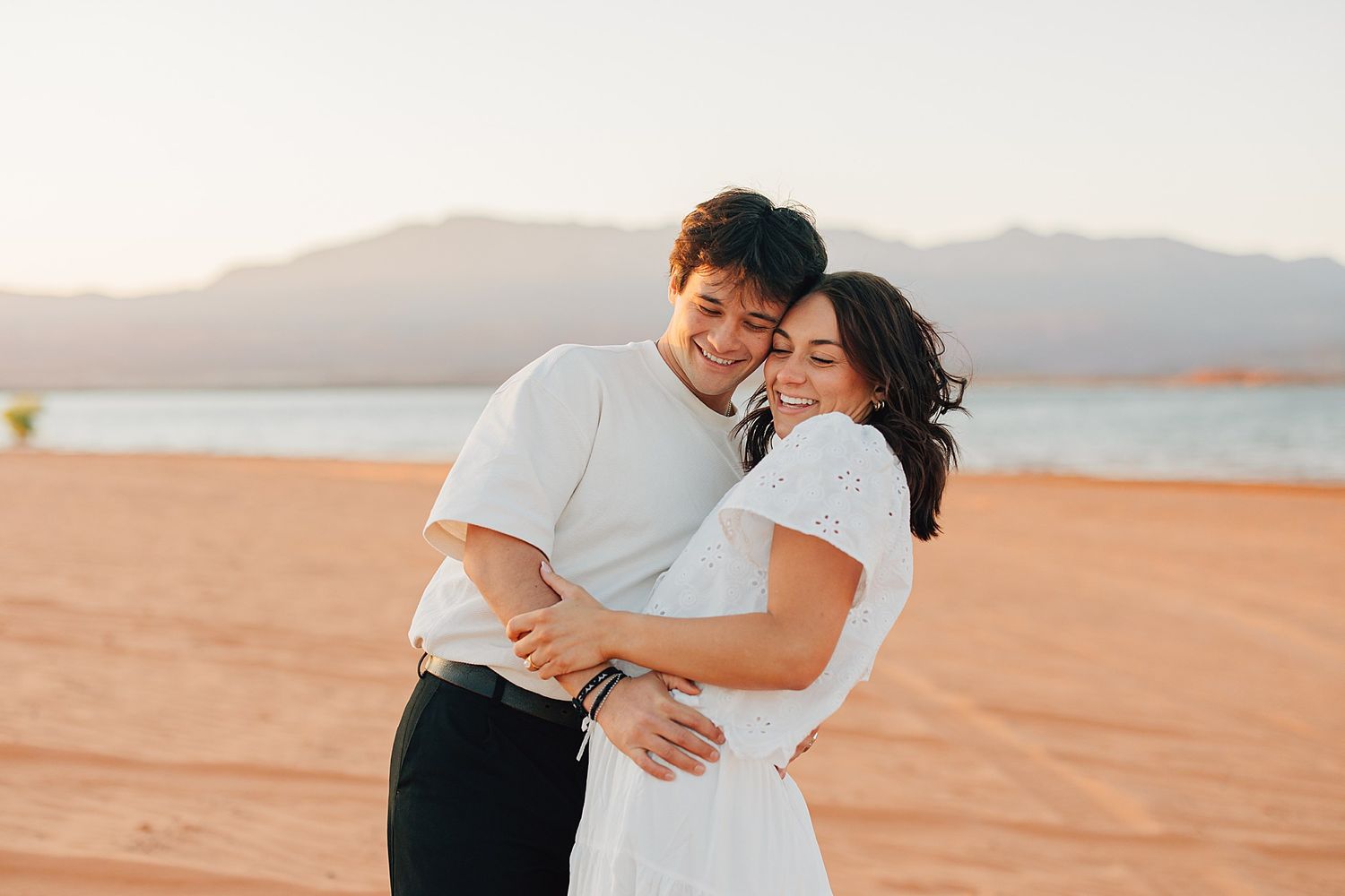 Couple embracing and smiling on a sandy beach at golden hour with mountains in the background.
