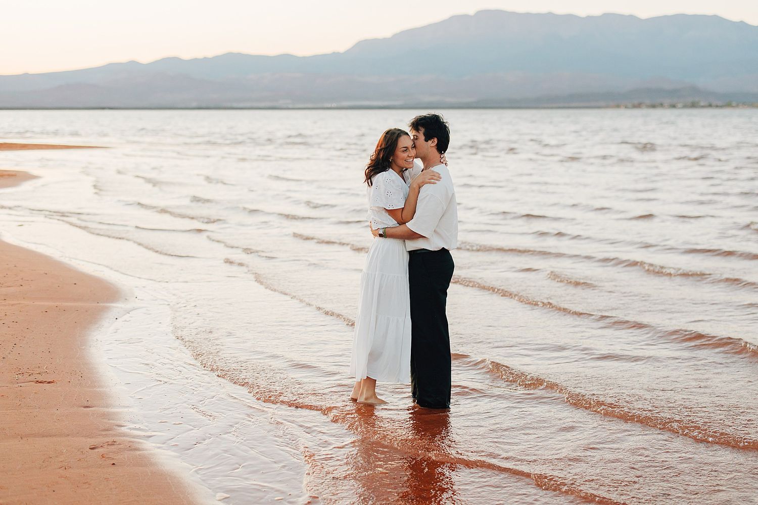 A romantic couple embraces barefoot on a serene beach at sunset, waves gently washing over their feet.