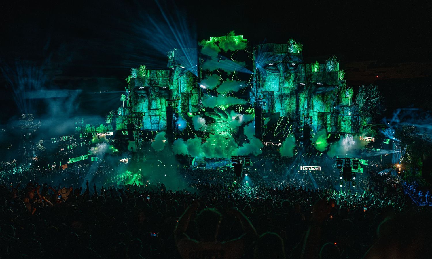 Massive festival stage illuminated in green light with a large crowd at night.