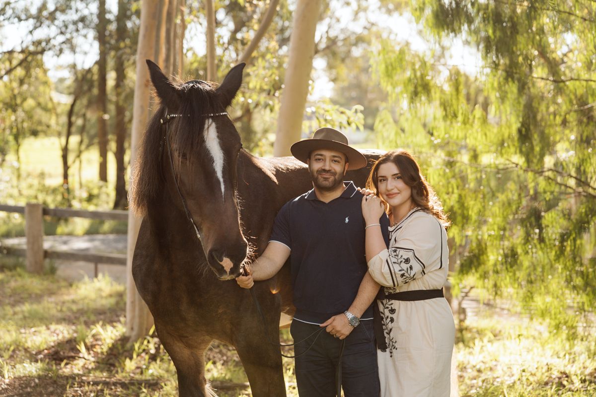 A couple in wide-brim hats pose with a dark horse in a sunny outdoor setting with trees and a wooden fence during a photoshoot in Sydney