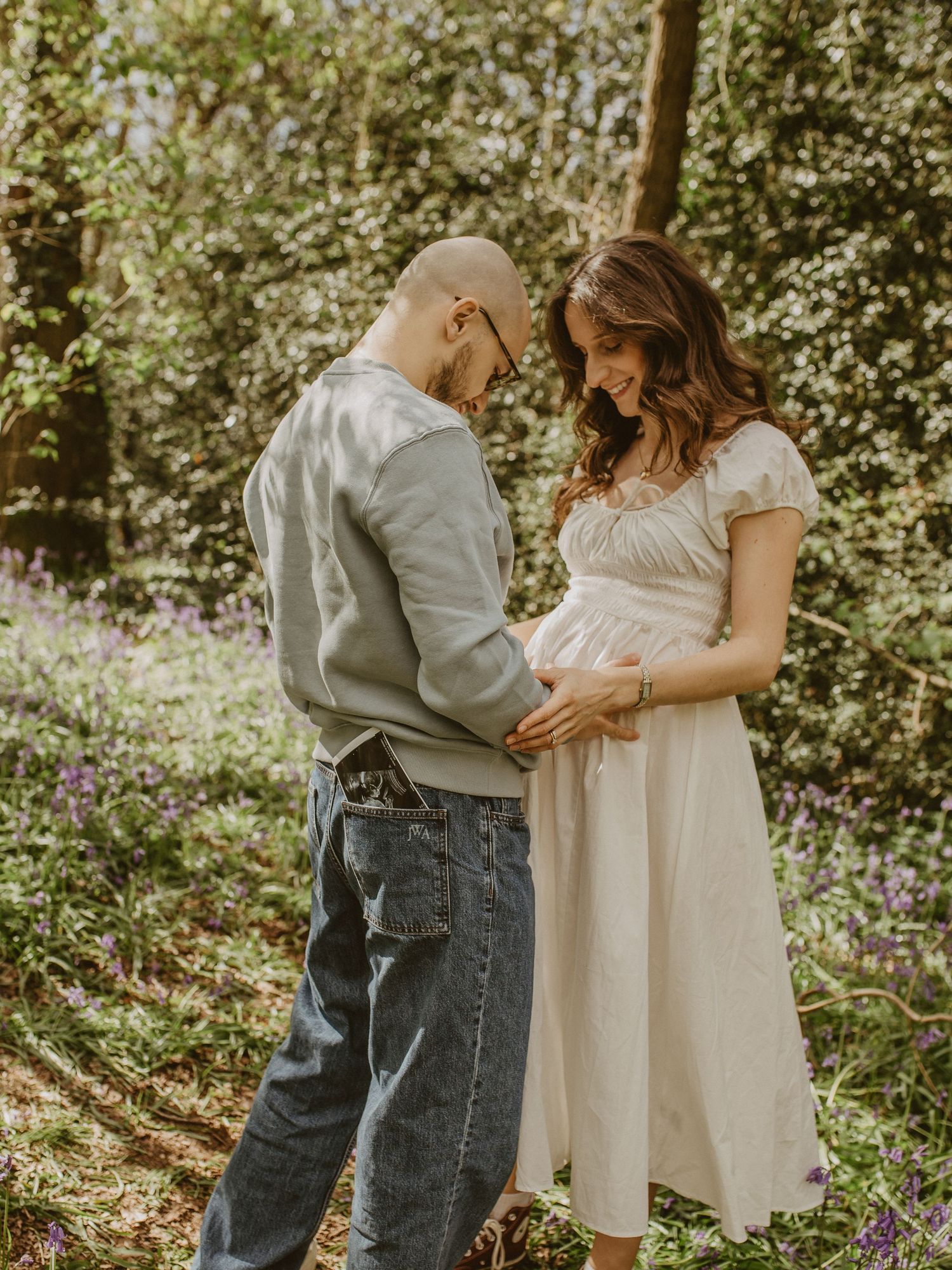 A couple tenderly cradles a baby bump in a sunlit bluebell woodland during a maternity photoshoot.