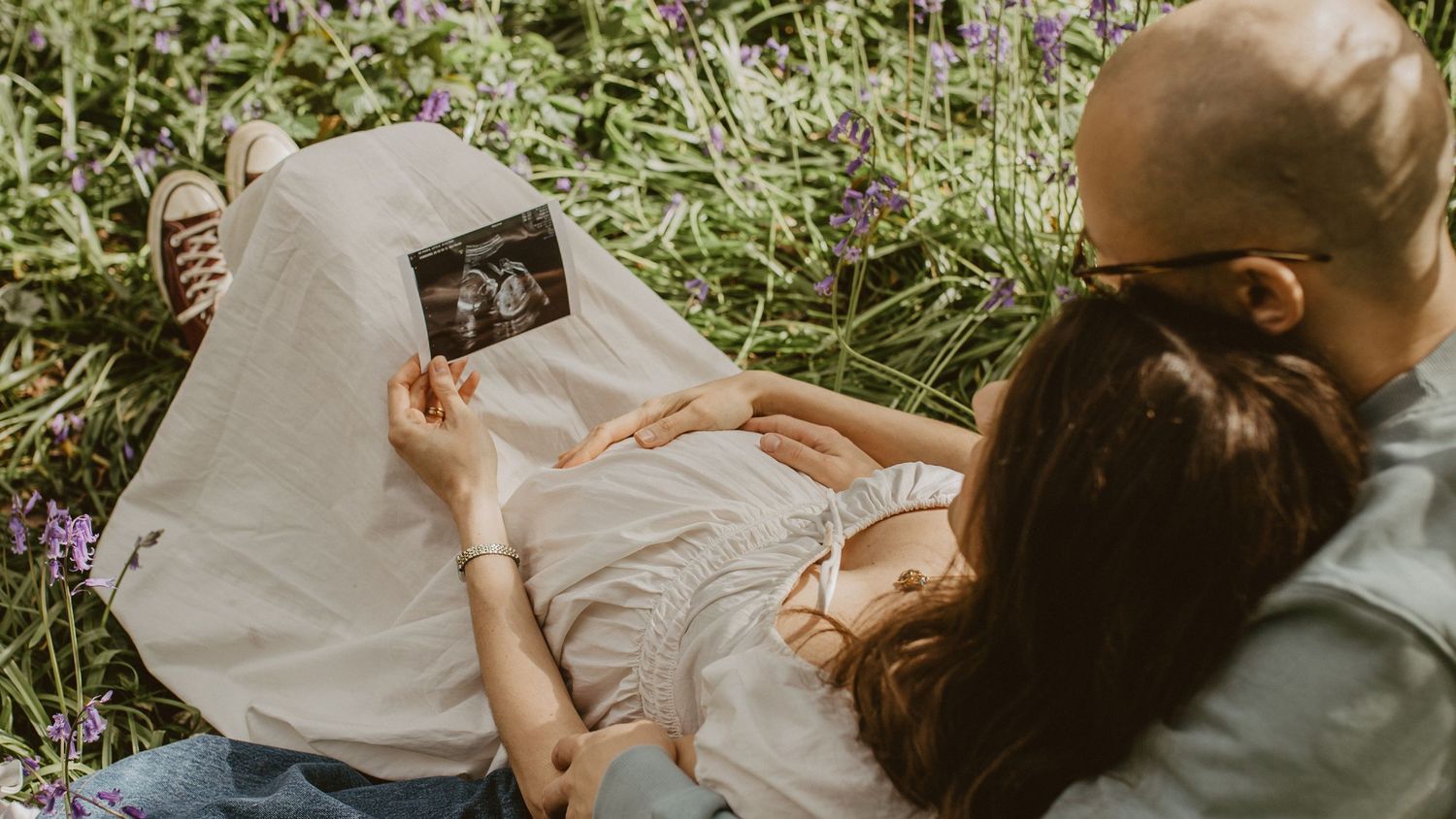 A couple sits in a flower field viewing their baby ultrasound photo together during a maternity announcement session.