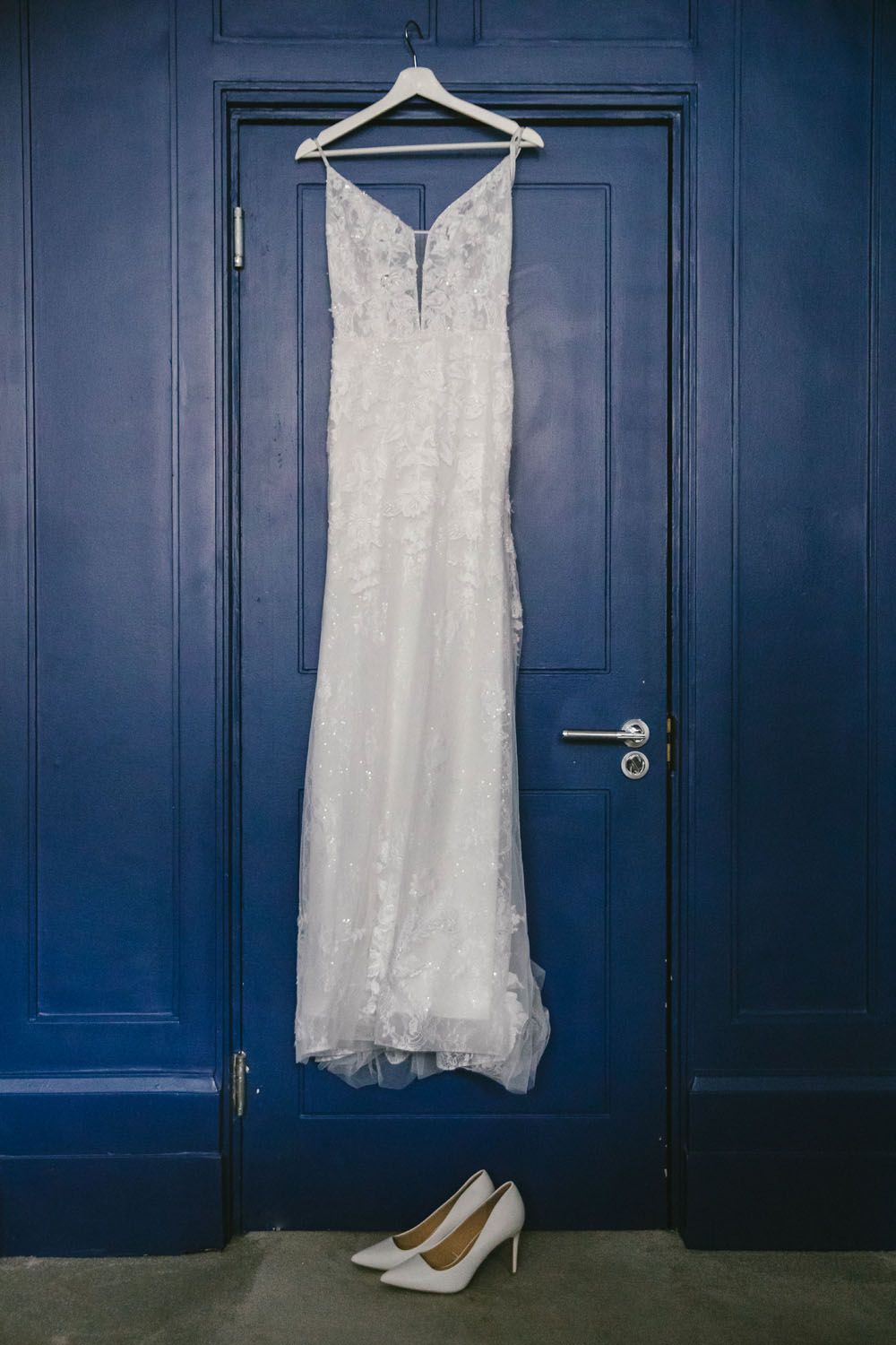 White lace wedding dress hanging on blue door with matching white heels below.