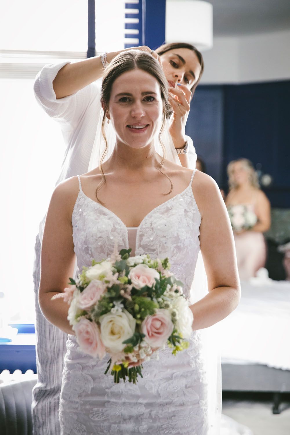 Bride in white lace gown holds pink bouquet while bridesmaid adjusts her veil before the wedding ceremony.