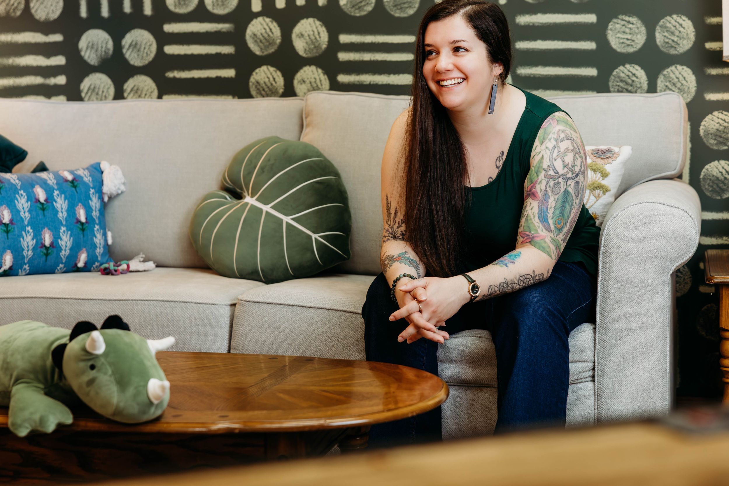 Smiling woman with long hair and tattoos sits relaxed on a white chair in a cozy, decorated living room.