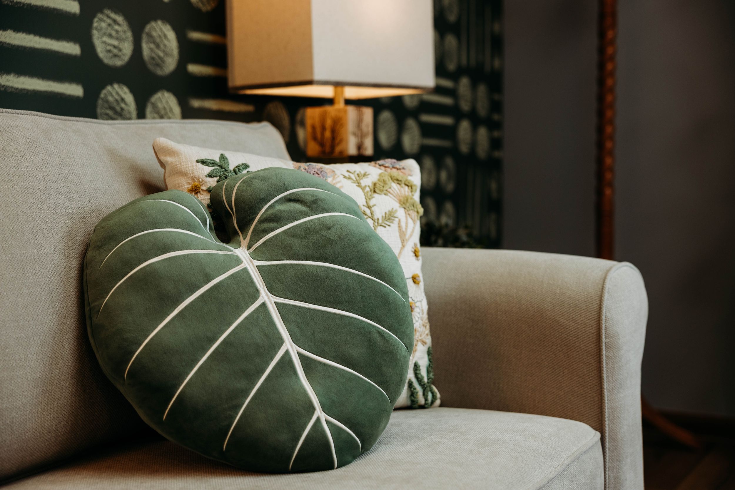 Green leaf-shaped velvet pillow on a light gray sofa with floral cushions and a lamp in the background.