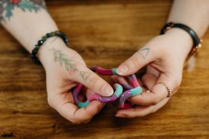 Tattooed hands kneading colorful pink and teal clay on a wooden surface, wearing bracelets and rings.