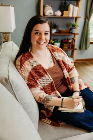 Smiling young woman with tattoos writing in notebook on couch, wearing striped cardigan in cozy home setting.