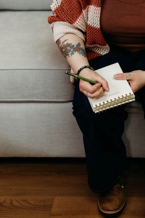 Person with floral tattoo writing in spiral notebook on couch, wearing striped sweater and brown shoes.