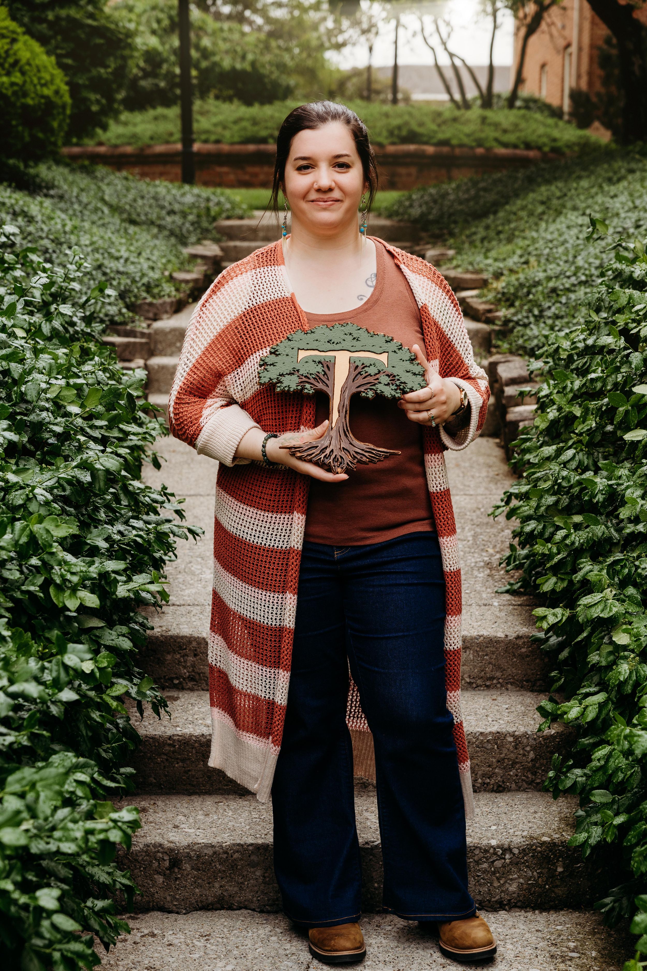 Woman in striped cardigan holds decorative bonsai tree sculpture on garden steps surrounded by lush greenery.