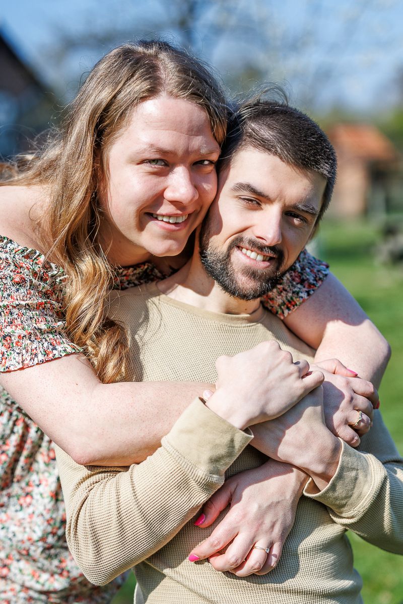 Happy couple embracing outdoors in spring, smiling woman hugging man from behind in a sunny garden setting.