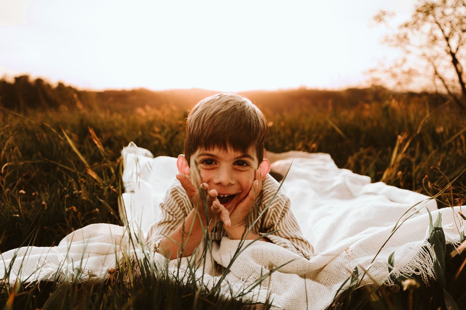 A joyful boy lying on a white blanket in a golden sunlit field, smiling with hands on cheeks.