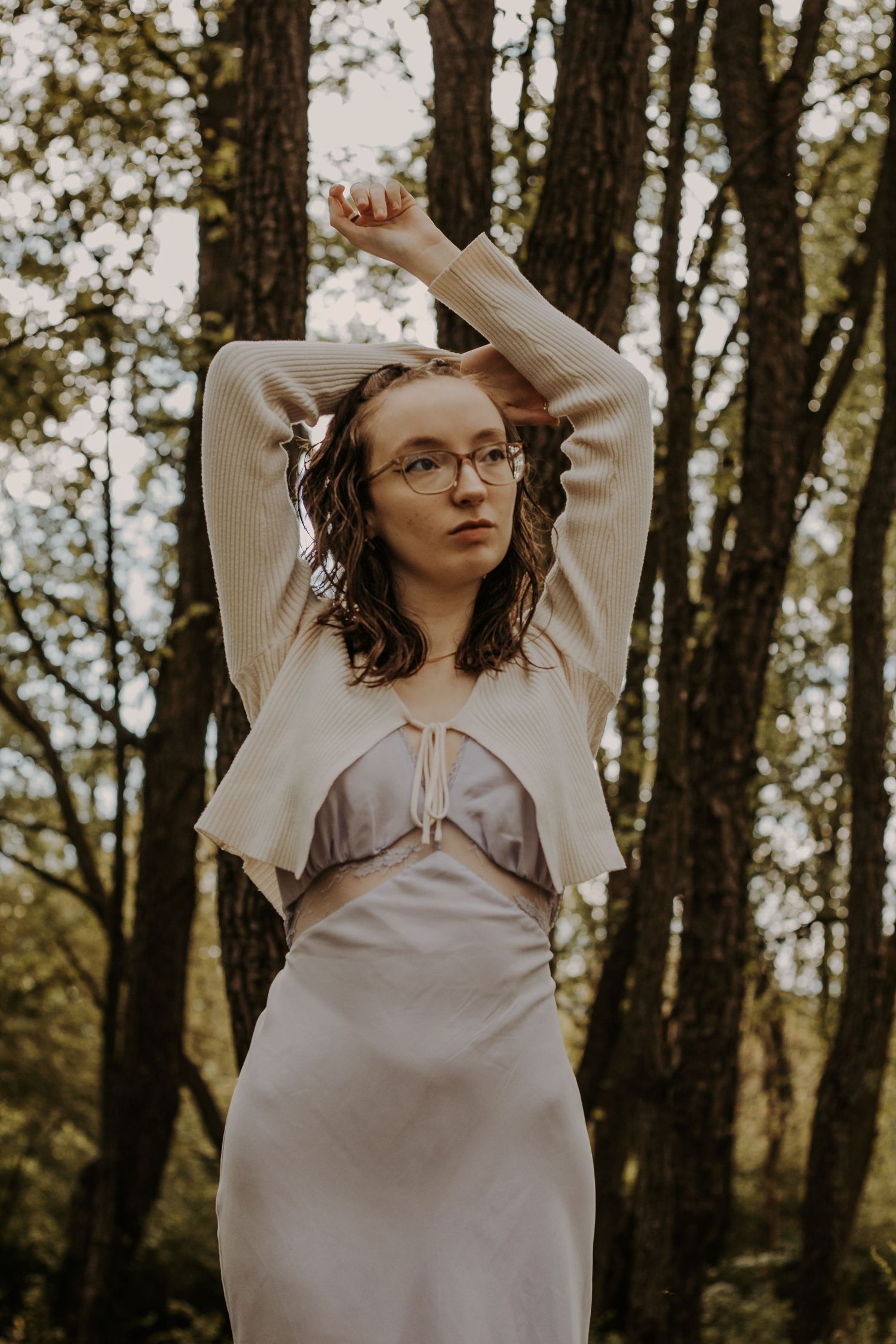 Young woman in cream knit cardigan and silver slip dress poses with arms raised in a forest setting.