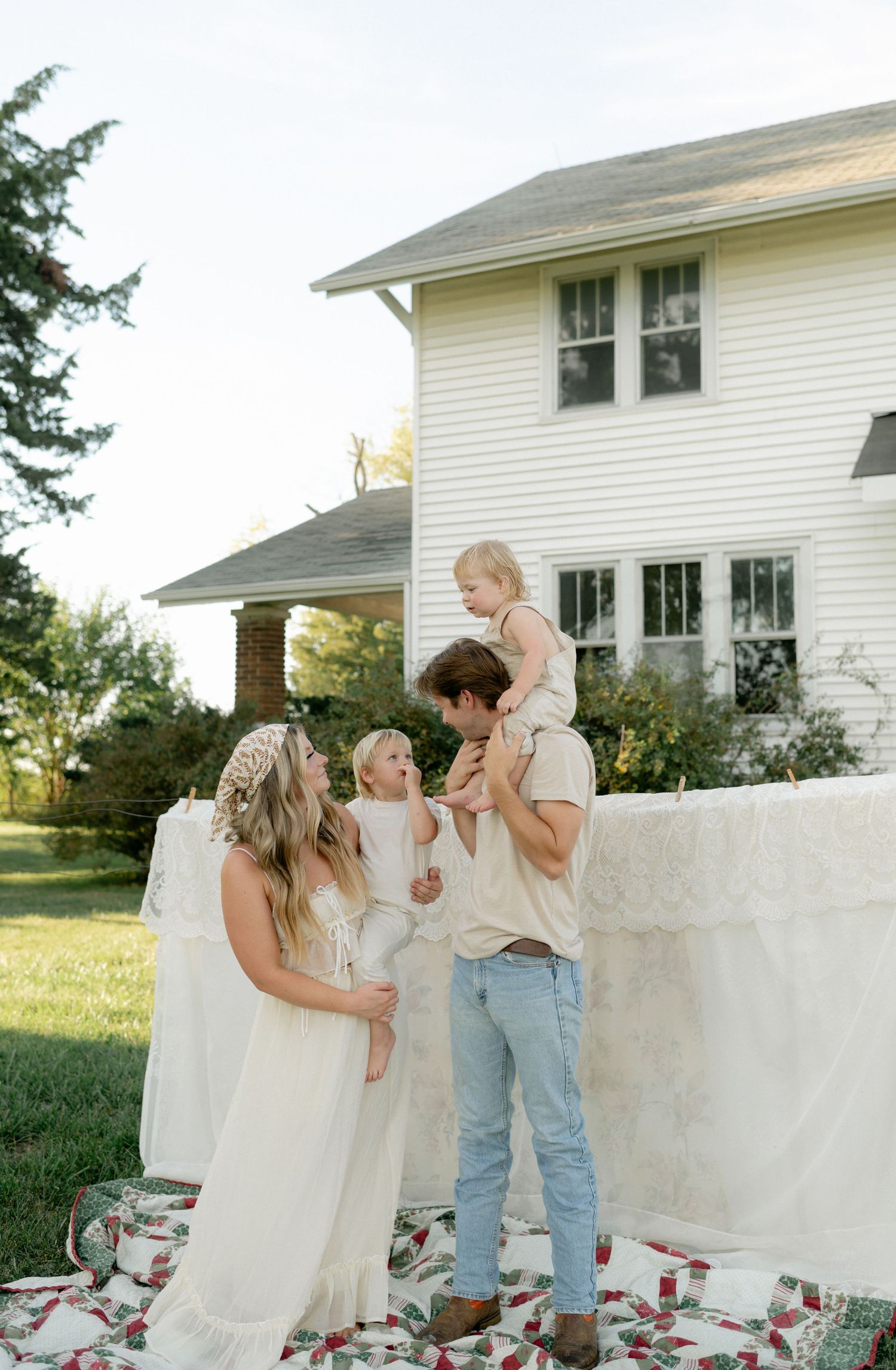 Boho family poses on patchwork quilt in front of white farmhouse during golden hour sunset.