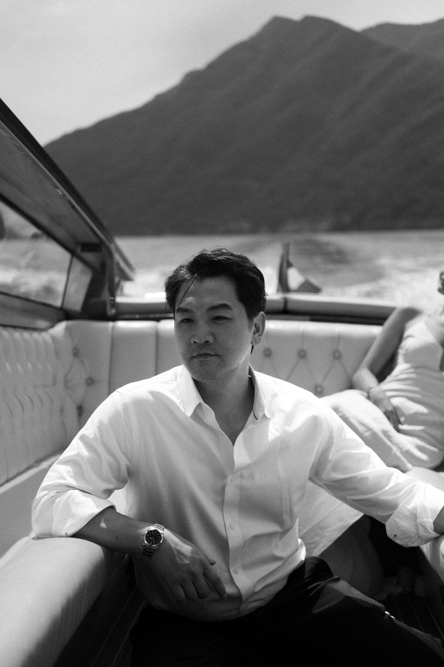 Black and white photo of a man in white shirt sitting on a luxury boat with mountain backdrop.