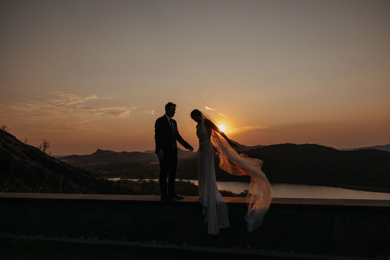 Silhouette of bride and groom holding hands at sunset with scenic water and mountains in background.