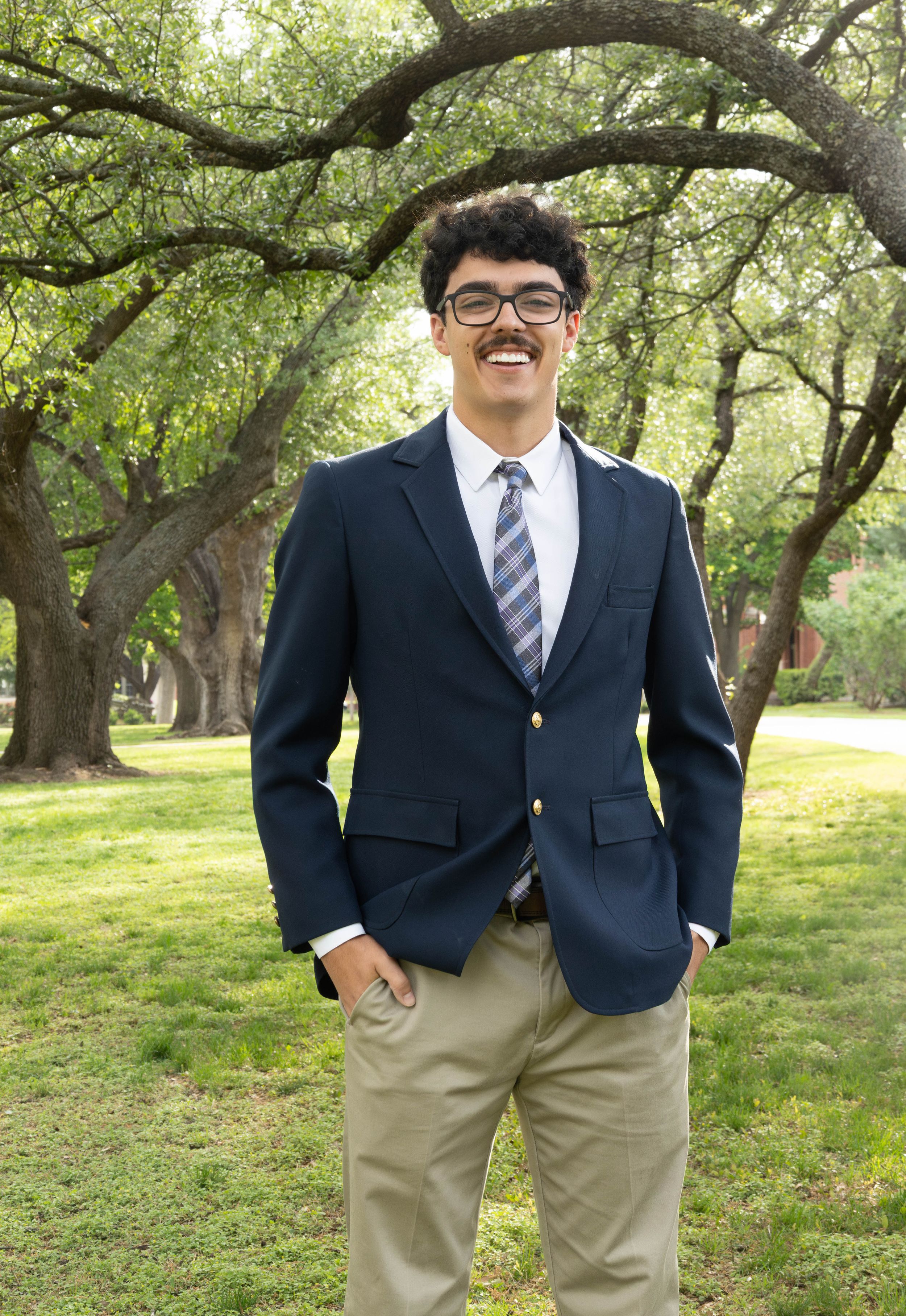 Young man in navy blazer, striped tie, and khakis smiling outdoors among green trees on a sunny day.