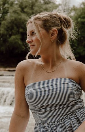 Woman in striped strapless top with gold necklace and earrings, smiling outdoors near a waterfall.