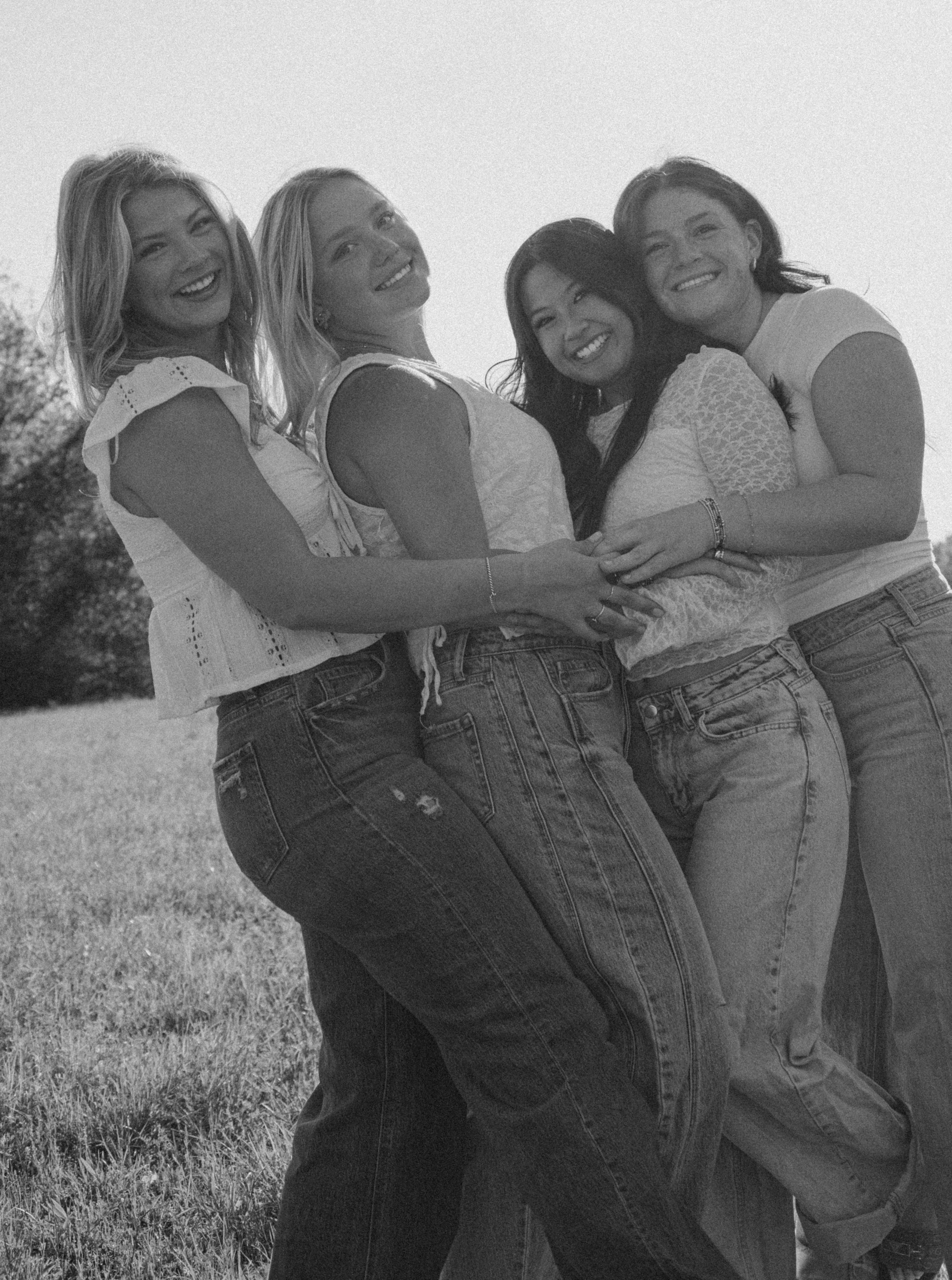 Four young women smiling and hugging outdoors in a black and white photo, wearing jeans and casual tops.