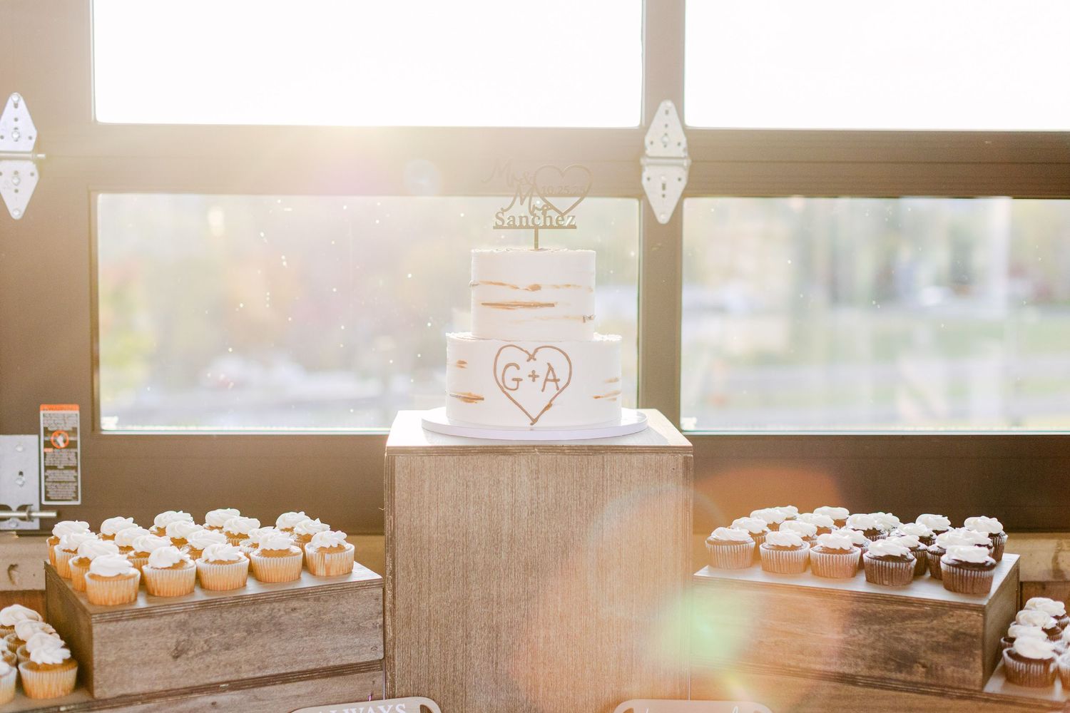 White wedding cake with Mr & Mrs Sanchez cake topper dated 10.25.25, displayed in warm sunlit venue.