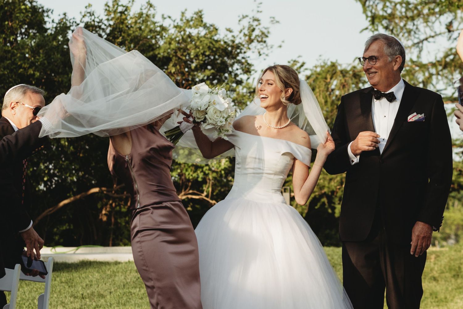 Bride in white ball gown laughing as veil blows in wind, flanked by family members at outdoor wedding ceremony.