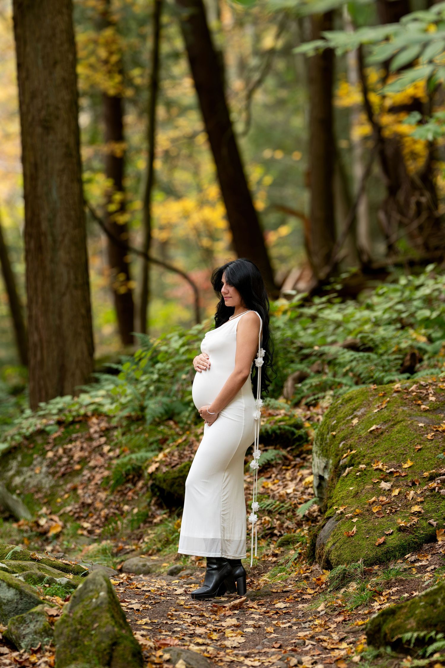 Pregnant woman in white dress and black boots standing on a forest path surrounded by autumn foliage and mossy rocks.
