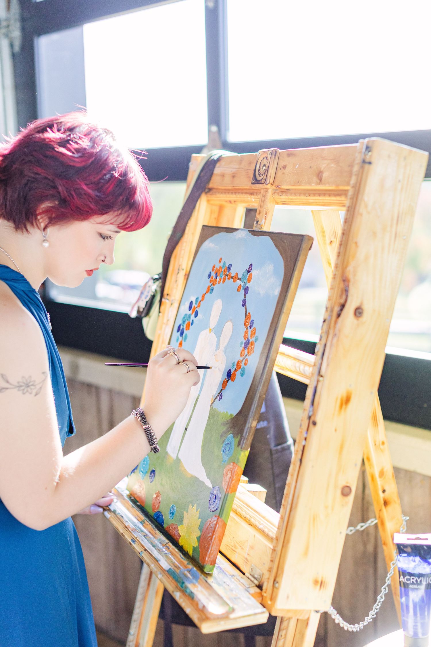 Red-haired woman in blue dress painting colorful artwork on wooden easel in bright studio.