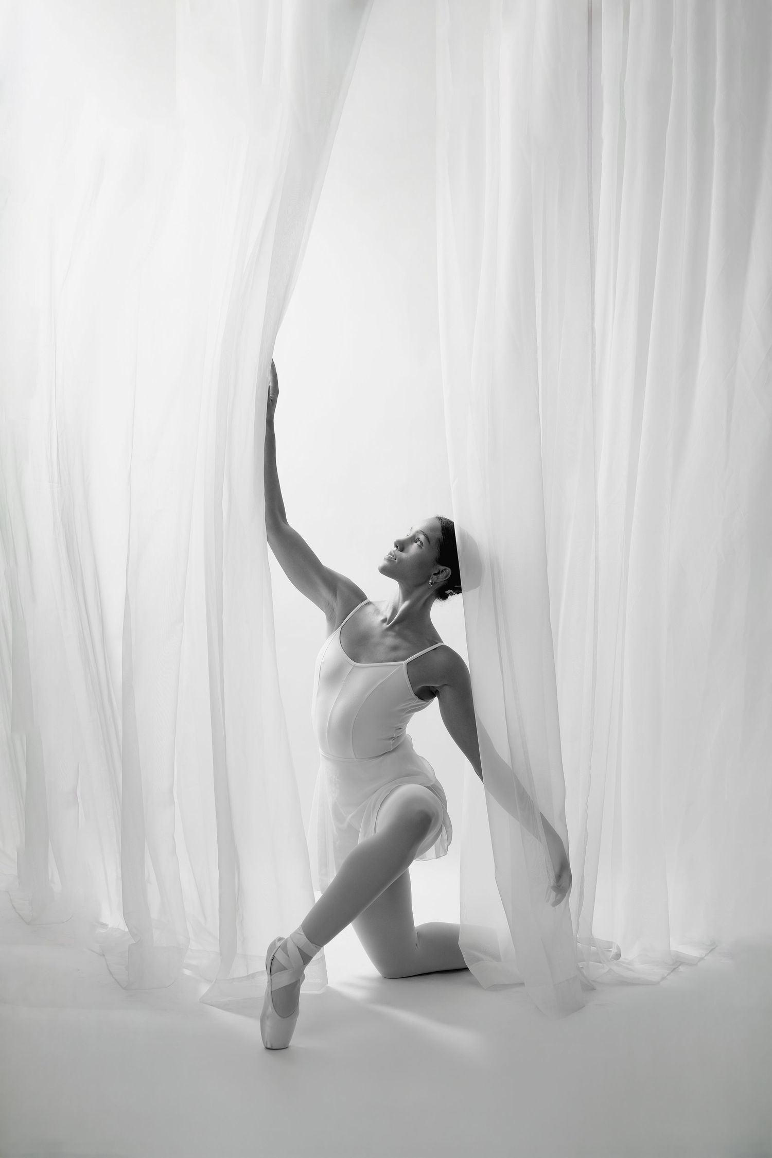 Black and white photo of a graceful ballerina in white leotard and pointe shoes posing elegantly with sheer curtains.