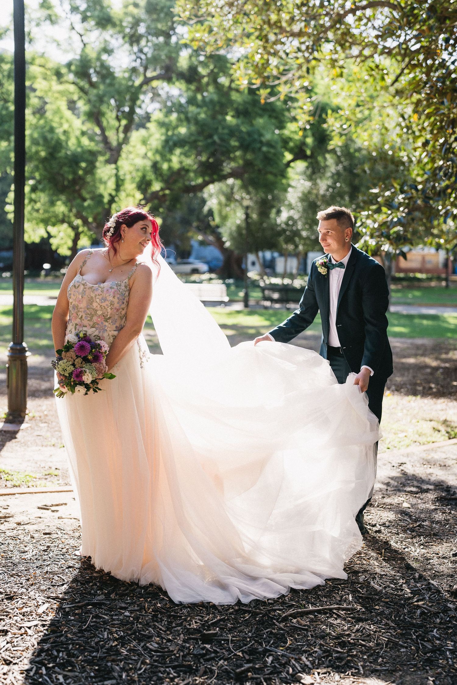 Groom lifts bride's flowing blush tulle gown as they walk through a sunlit park on their wedding day.