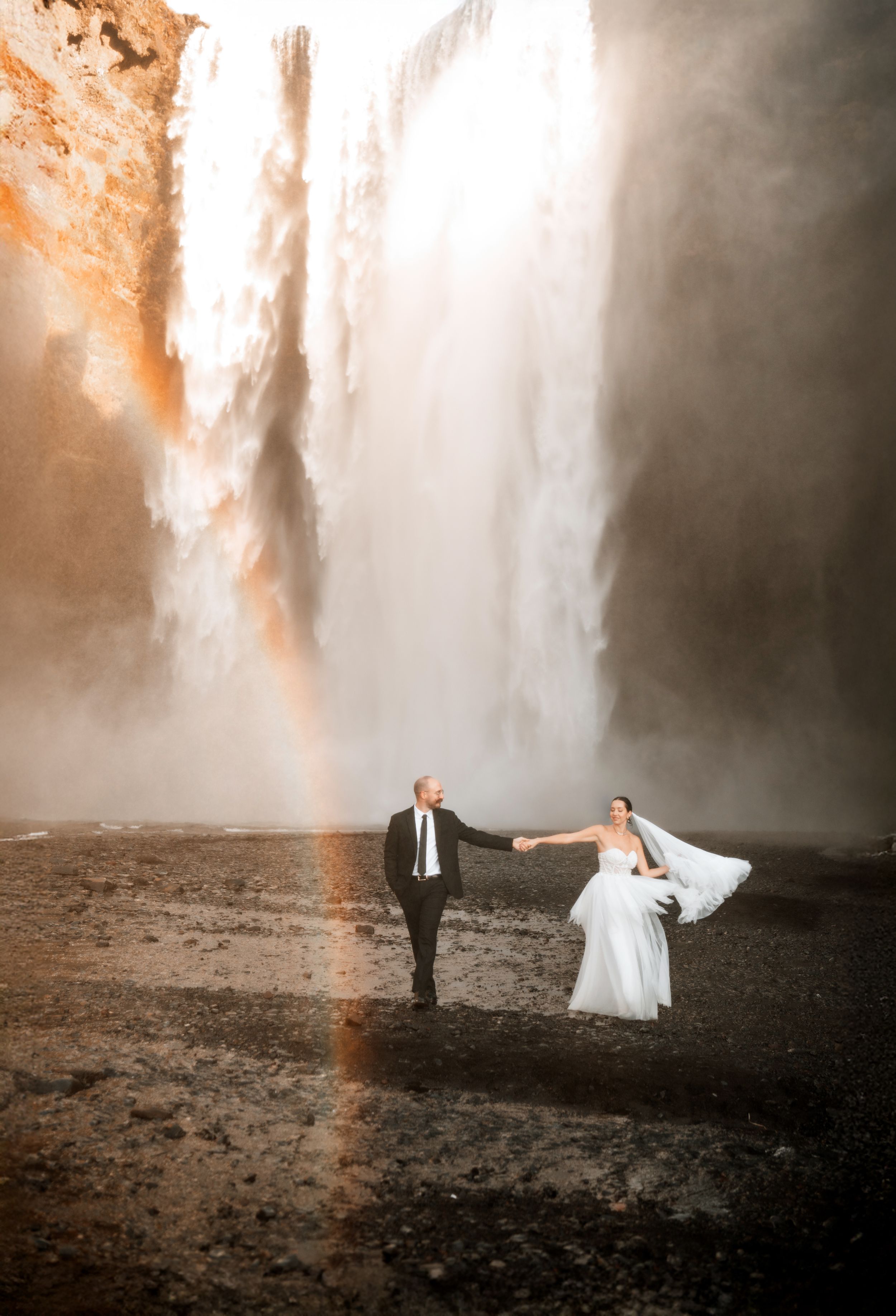 Bride and groom hold hands walking before a massive waterfall with a rainbow in misty light.
