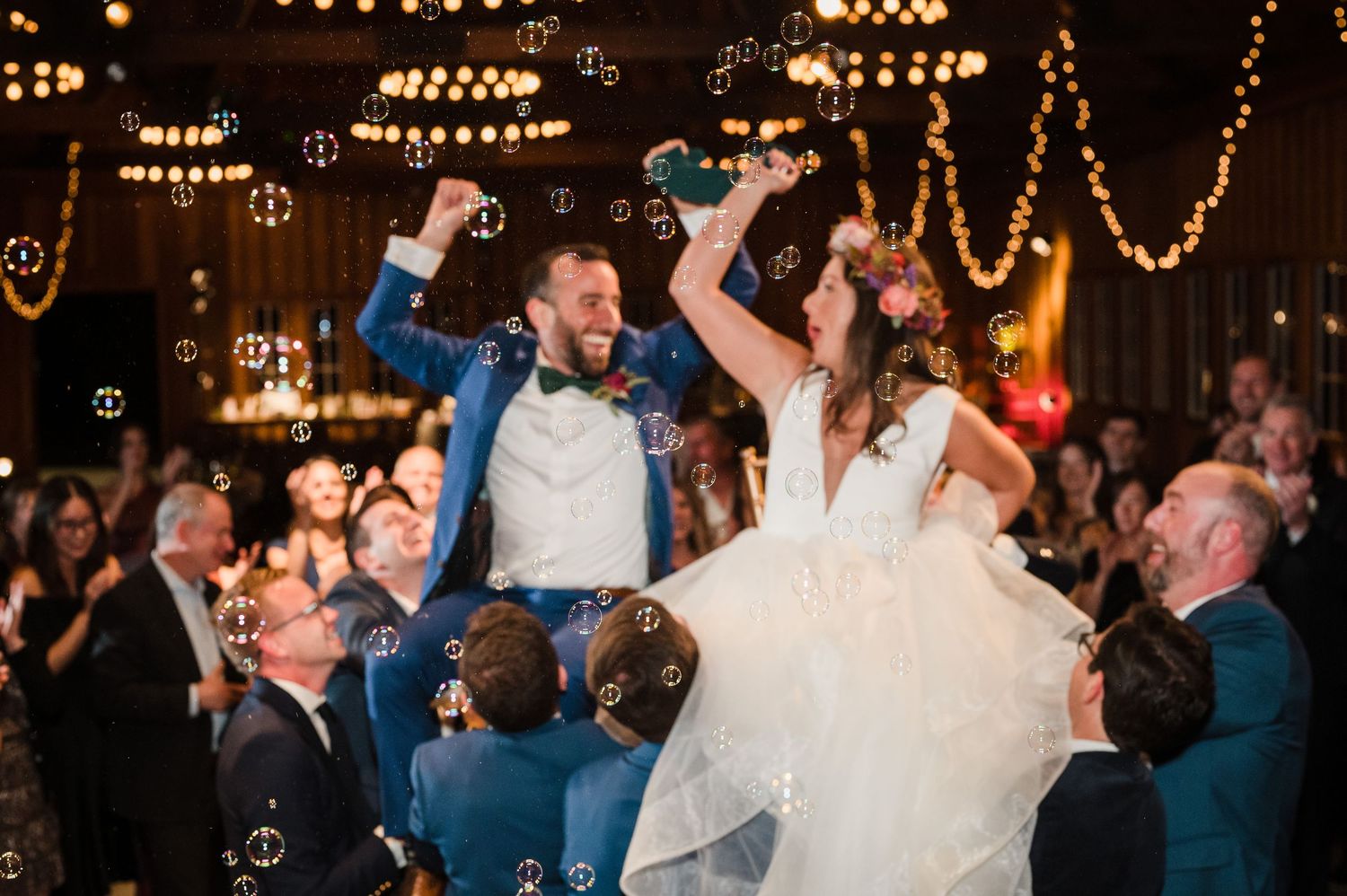 Joyful bride and groom lifted by guests at wedding reception with bubbles and string lights.