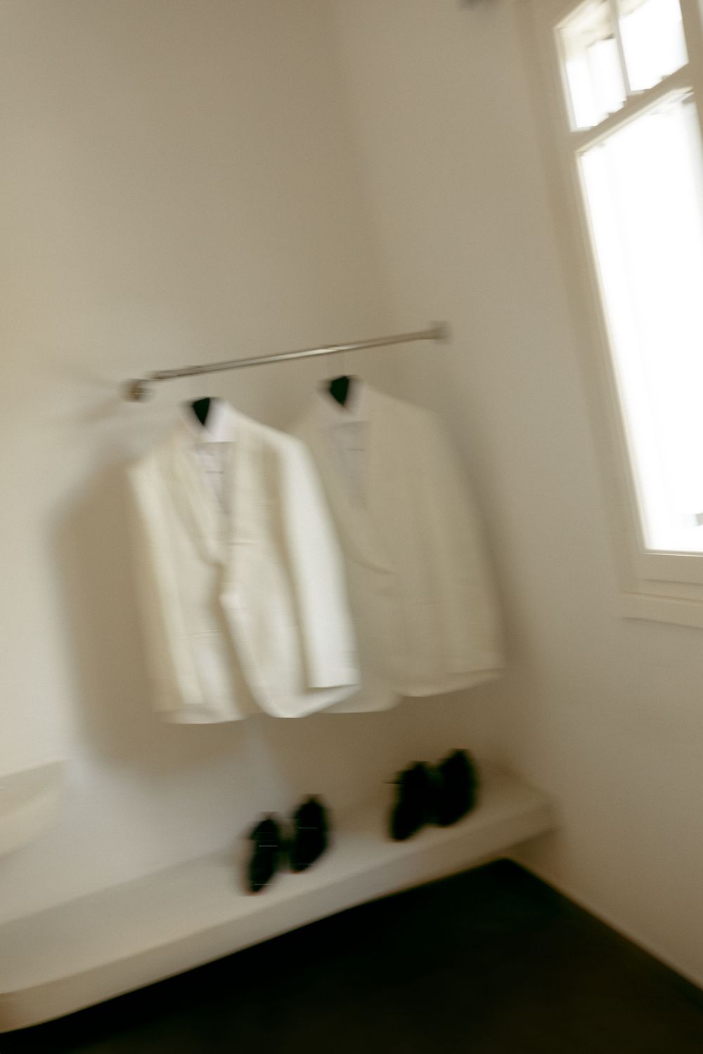 Two white tuxedo jackets hanging on rack with black dress shoes below in bright room.
