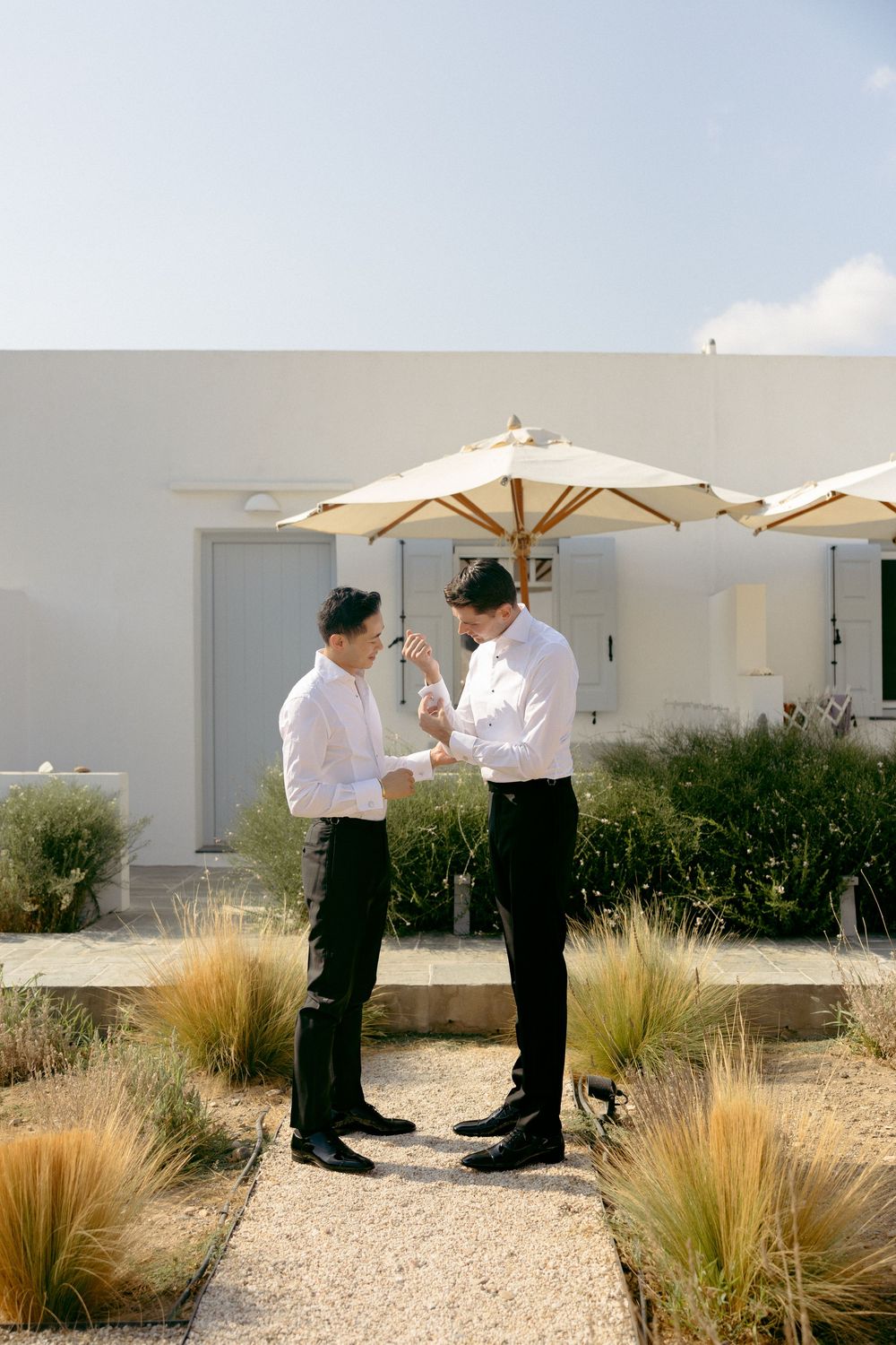 Two grooms in white shirts helping each other prepare in sunny Mediterranean villa garden.