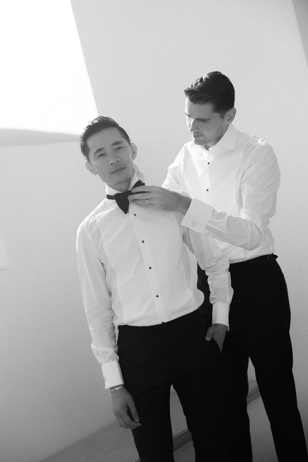 Black and white photo of two grooms in white shirts as one adjusts the others bow tie indoors.