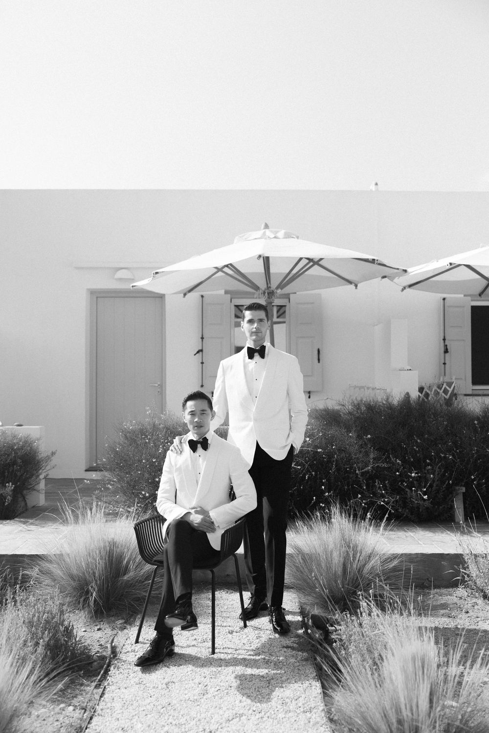 Black and white portrait of two grooms in white tuxedos posing outside a Mediterranean villa.