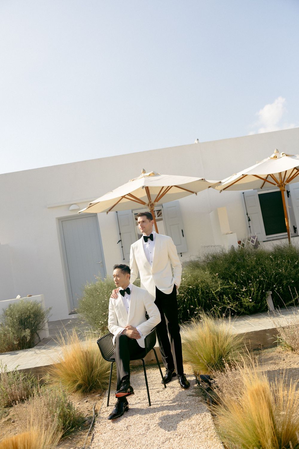 Two grooms in white tuxedos posing outdoors near umbrellas and white walls.
