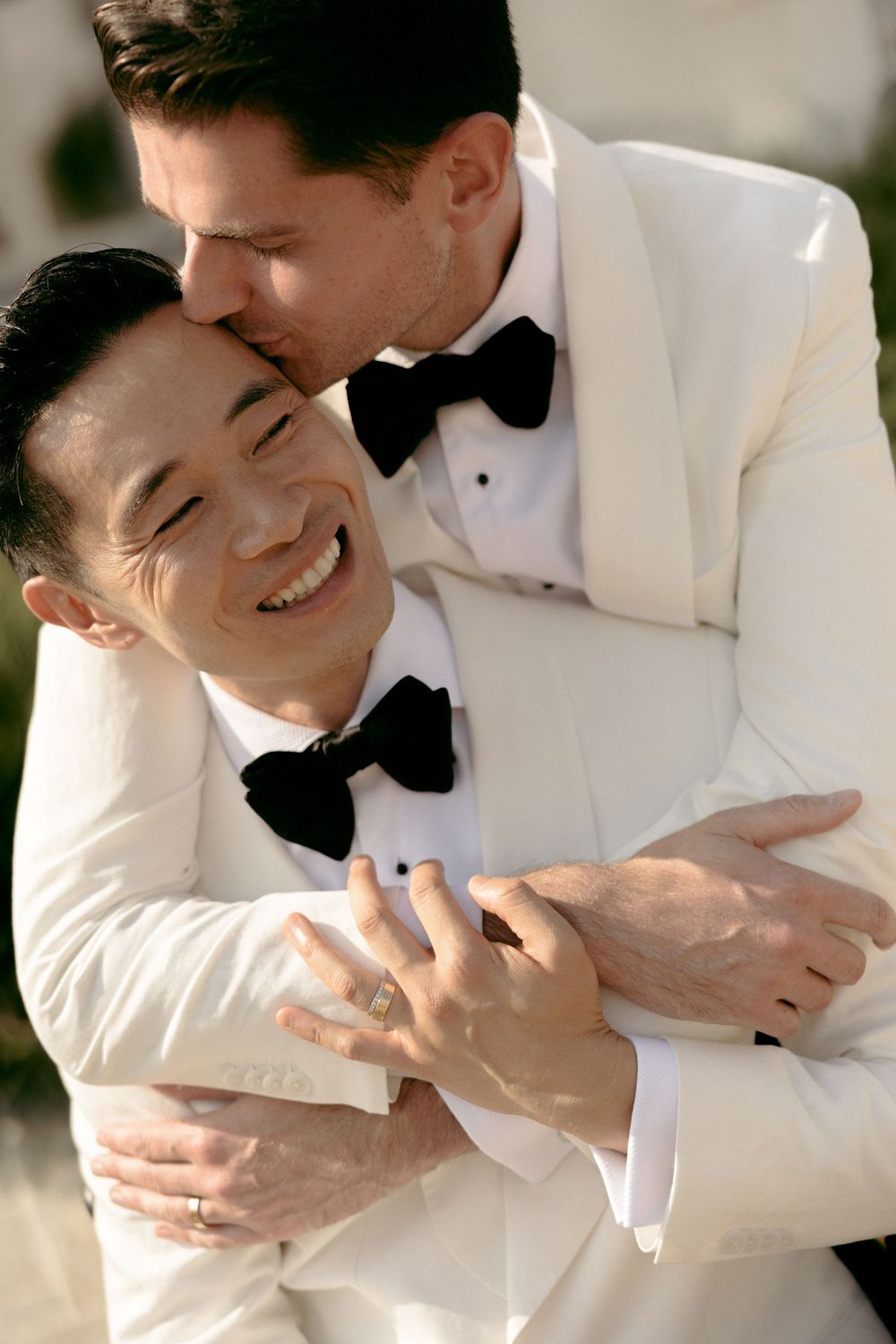 Groom in white tuxedo embraces and kisses his smiling partner at their wedding ceremony.