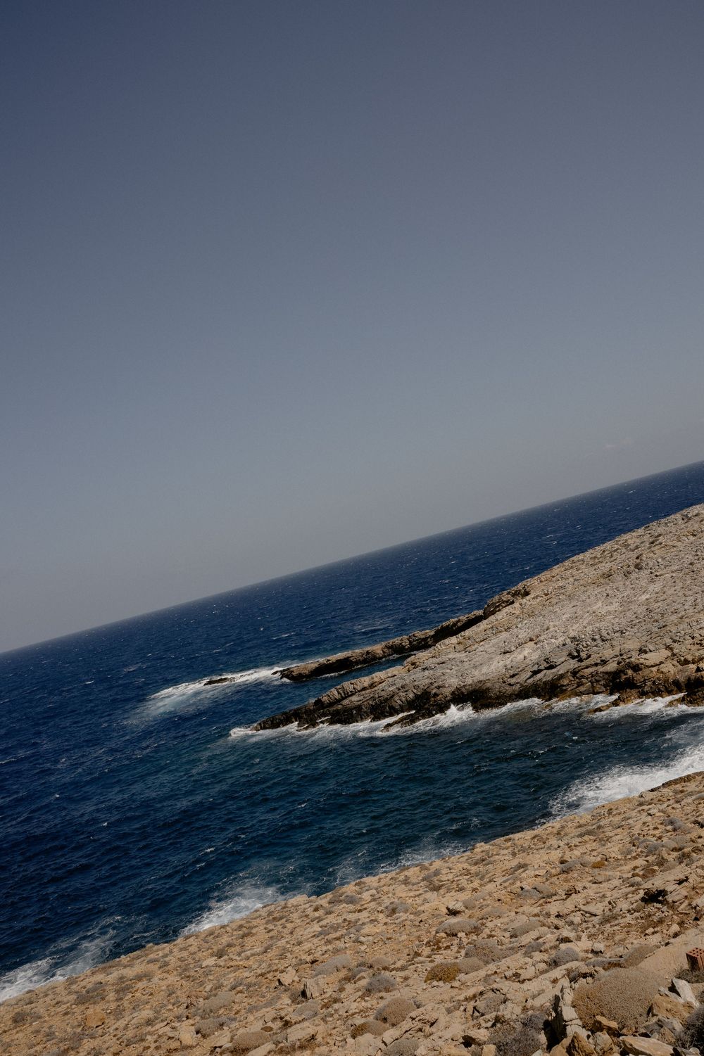 Rocky coastal cliffside overlooking deep blue Mediterranean sea waters under a clear hazy sky.