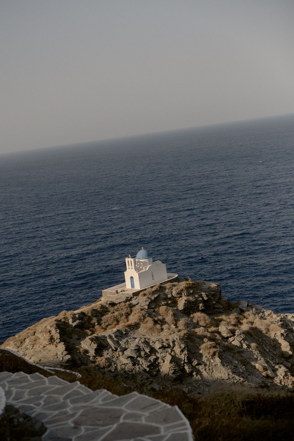 White Greek Orthodox chapel with blue dome perched on rocky cliff overlooking deep blue Mediterranean Sea.