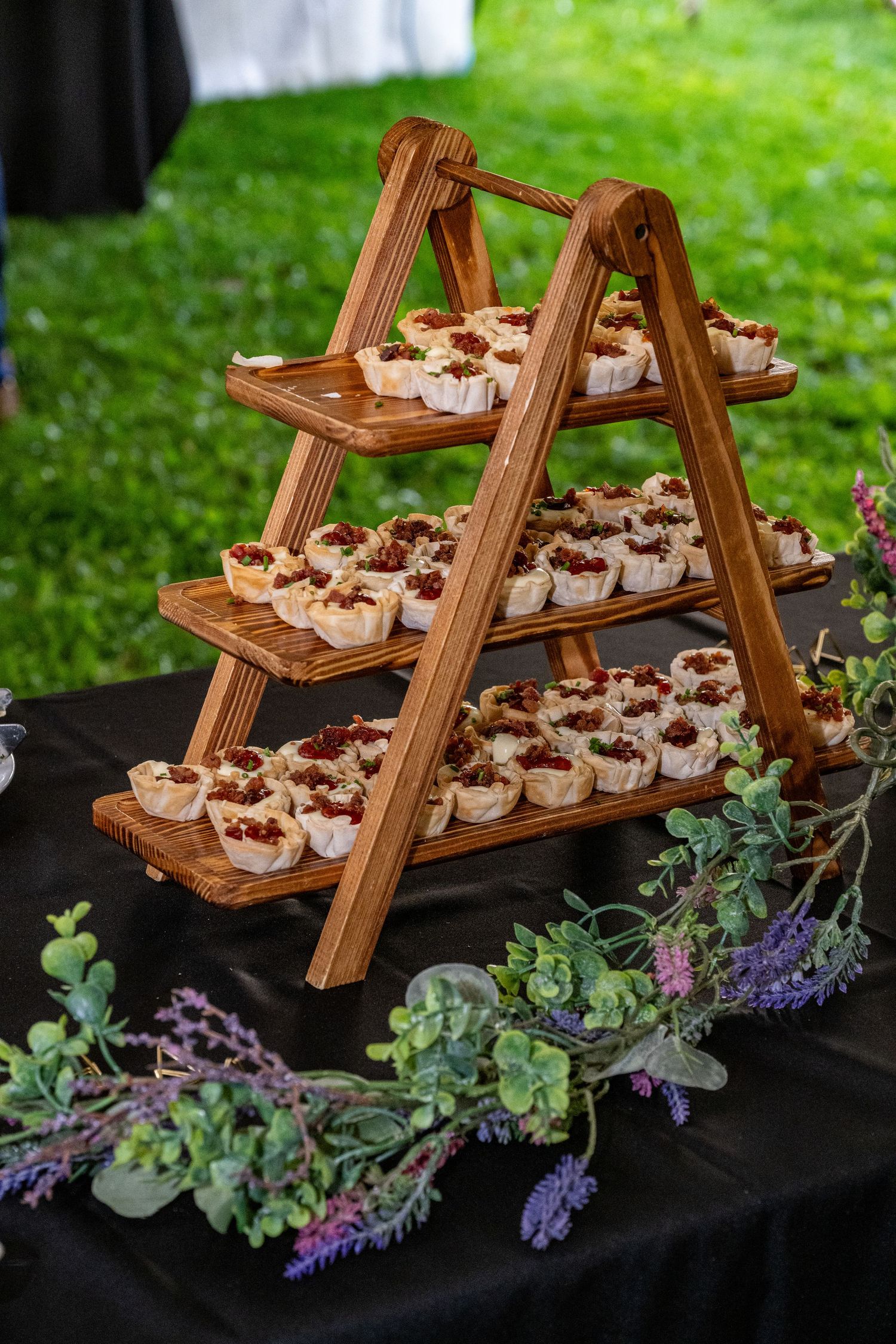 Three-tier wooden ladder display stand holding mini dessert tarts topped with berries on a black tablecloth with floral decor.