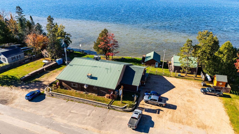 Aerial view of a lakeside log cabin with green metal roof, surrounded by trees and waterfront access on a calm lake.
