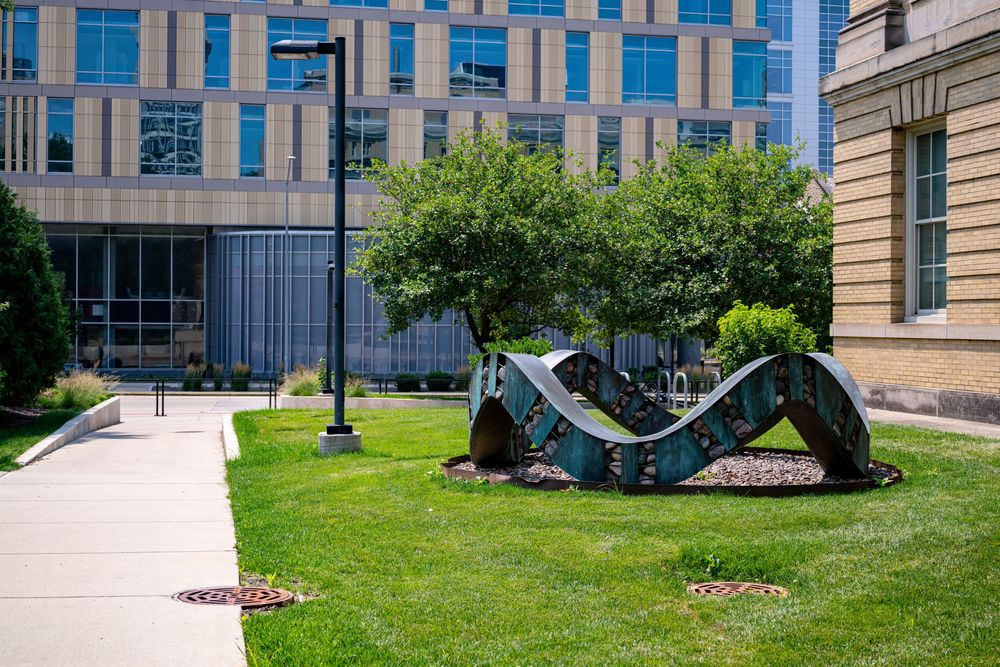 Colorful wave-shaped metal sculpture on green lawn between modern urban buildings on a sunny day.