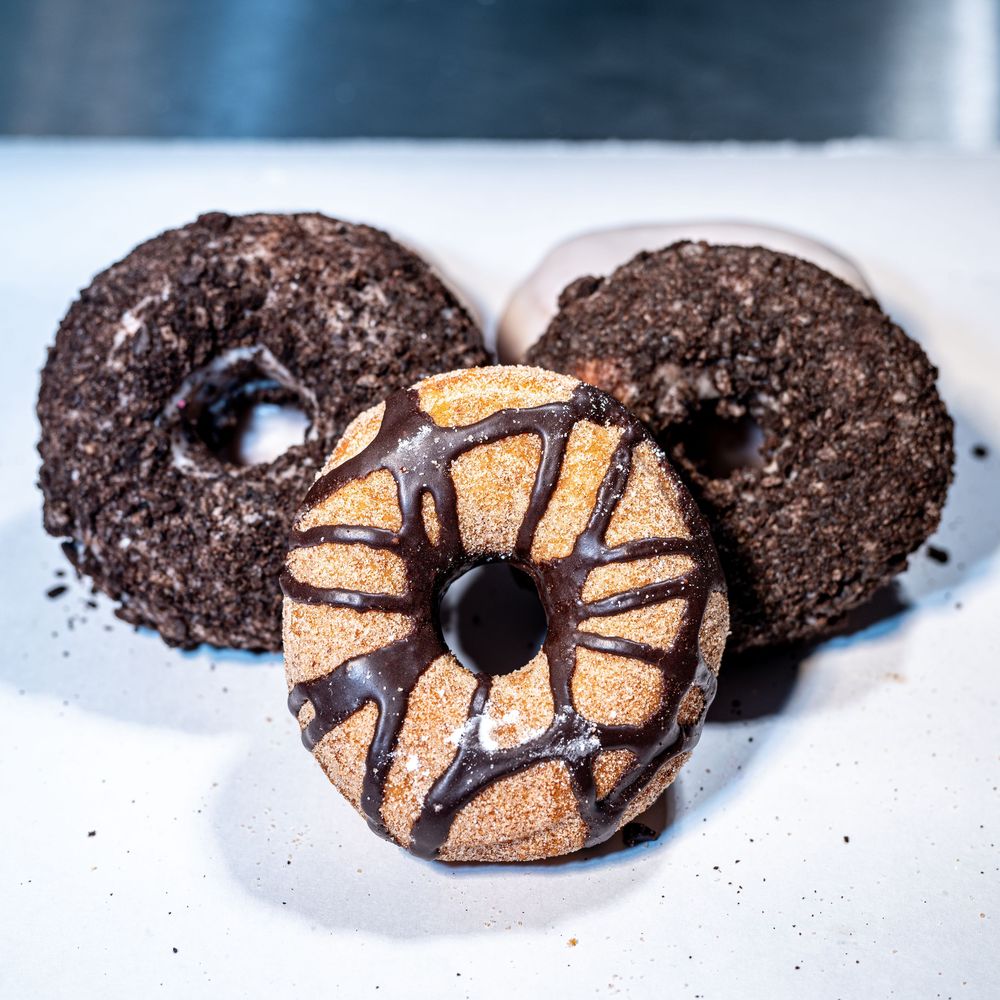 Three chocolate donuts on a white surface, two covered in dark crumbs and one with chocolate drizzle and cinnamon sugar.
