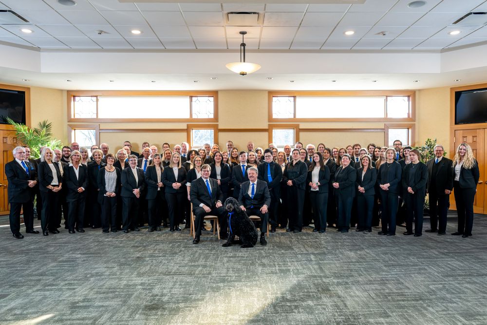 Large group of professionals in dark business attire posing together in a modern conference room with a dog in front.