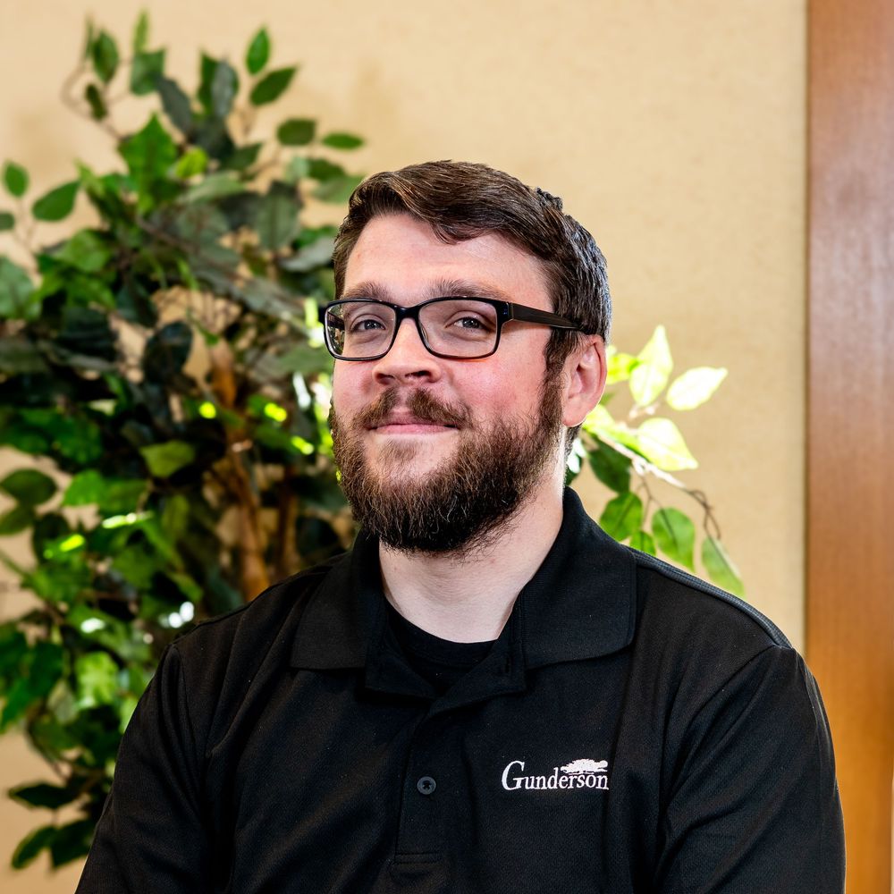 A smiling bearded man wearing black-rimmed glasses and a Gunderson polo shirt stands near a green plant.