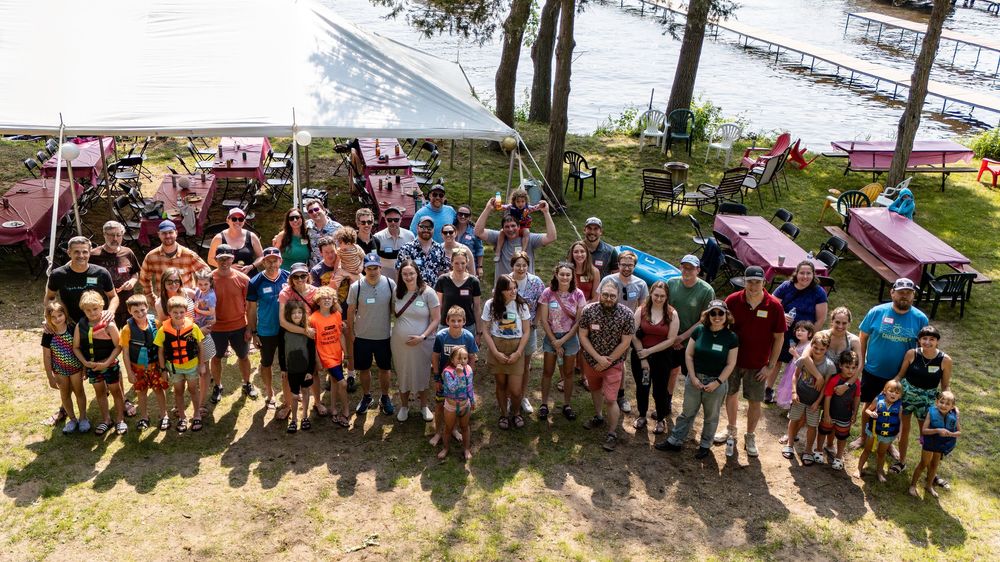 Large group of adults and children posing outdoors near a lake with colorful wooden structures on a sunny day.