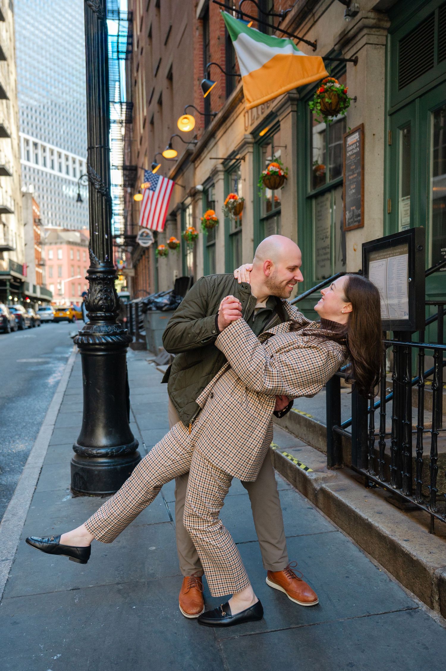 Couple dancing on a NYC street outside an Irish pub, man dipping woman in matching plaid suits with American flag backdrop.