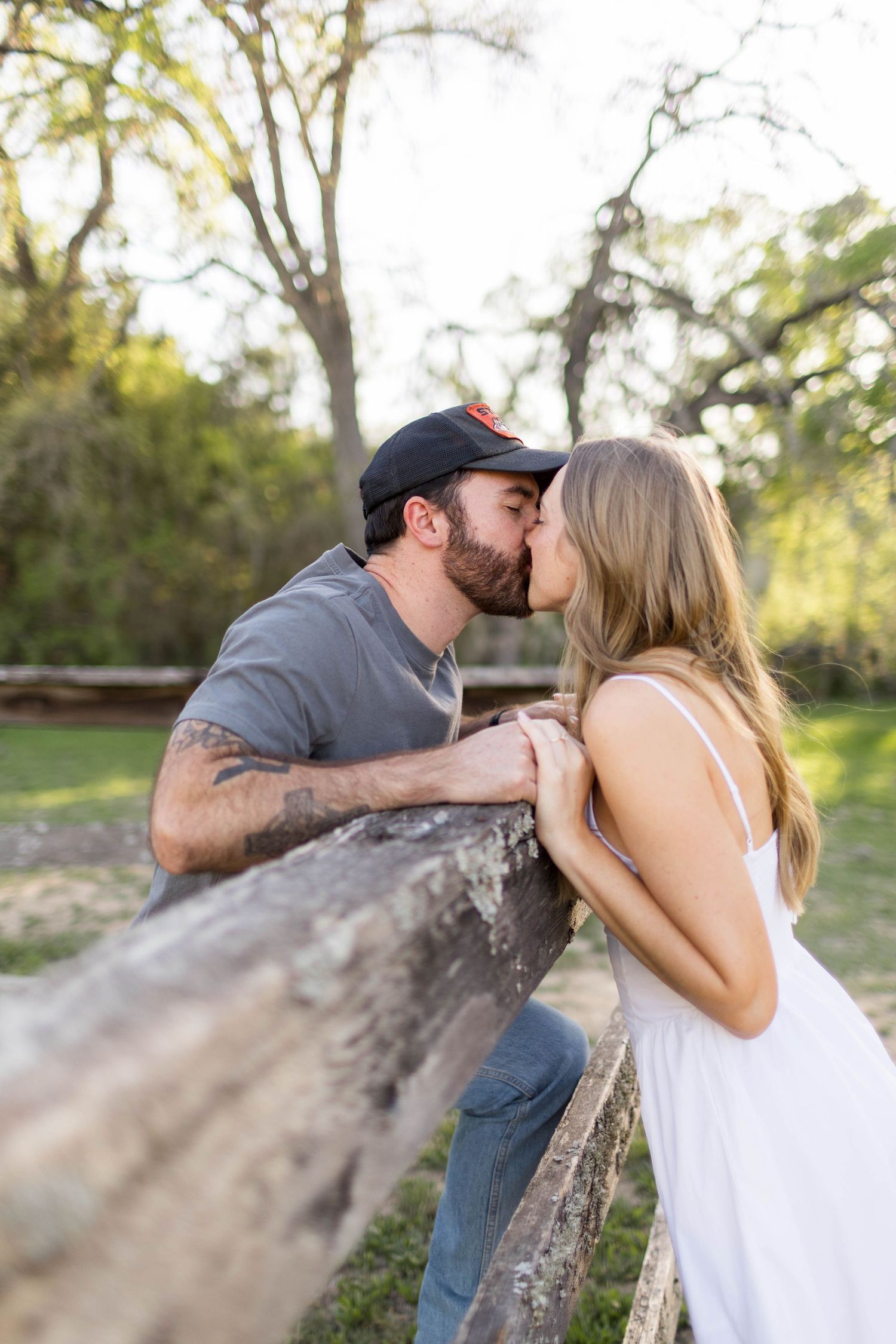 A couple shares a romantic kiss over a rustic wooden fence in a lush outdoor park setting at Commons Ranch in Austin, Texas.