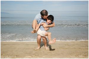 Man playfully lifting a child on a sandy beach with ocean waves in the background on a sunny day.